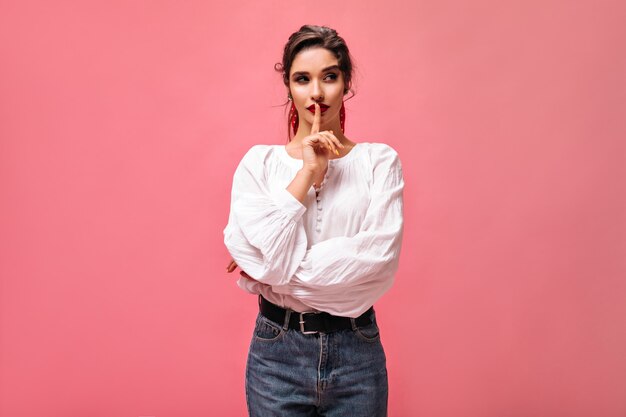 Mujer pensativa en poses de blusa blanca sobre fondo rosa. Linda chica con lápiz labial rojo con cabello oscuro en jeans con cinturón ancho mira a un lado.