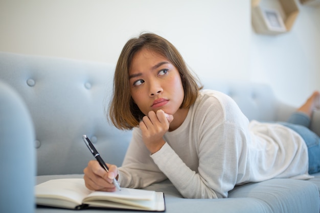 Mujer pensativa que hace notas en cuaderno y que miente en el sofá en casa