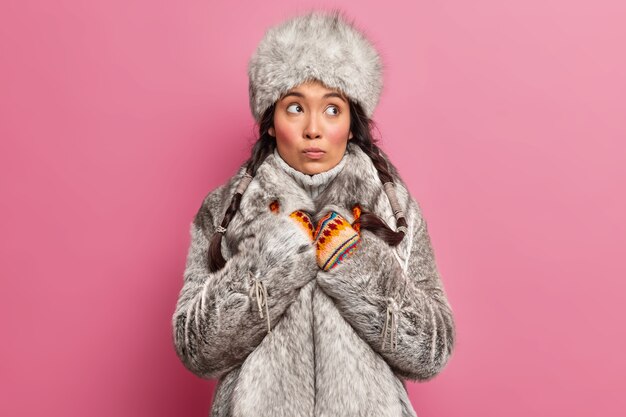 Mujer pensativa residente del lejano norte viste ropa abrigada de invierno mira hacia otro lado vive en la tundra plantea contra la pared rosa