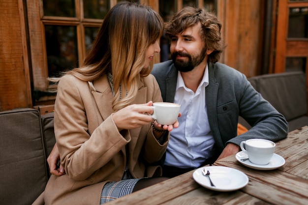 Mujer pensativa romántica con largos pelos ondulados abrazando a su marido con barba. Elegante pareja sentada en la cafetería con capuchino caliente.