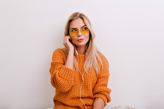 Mujer pensativa rubia con ojos grandes escuchando música mientras está sentada en su habitación de luz