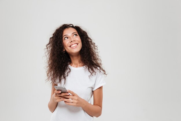 Mujer pensativa sonriente que sostiene smartphone en manos y que mira para arriba sobre fondo gris