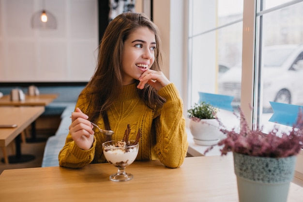 Mujer pensativa en suéter de punto mirando la ventana durante el descanso en la cafetería en un día frío. Retrato interior de mujer romántica en camisa amarilla disfrutando de un café en el restaurante.
