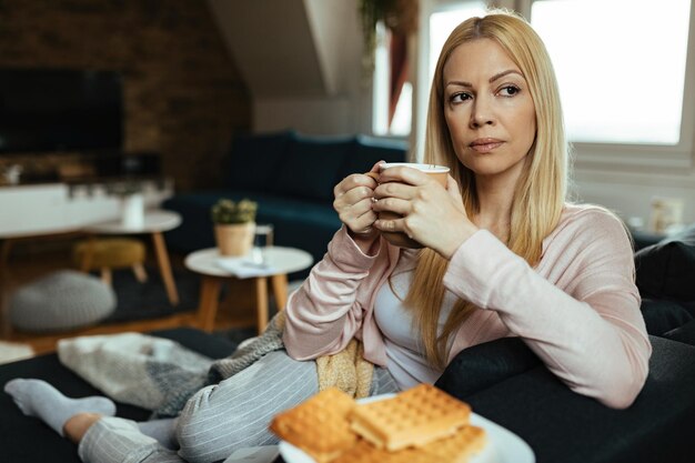 Mujer pensativa tomando café mientras se sienta en el sofá y piensa en algo en casa