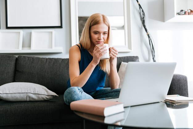 Mujer pensativa tomando café y usando la computadora portátil en casa