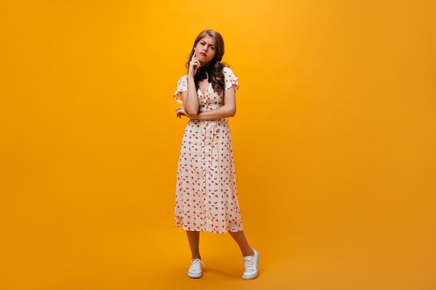 Mujer pensativa en vestido midi posa sobre fondo naranja. Chica triste con peinado rizado en ropa de moda fresca y zapatillas de deporte mirando a cámara.