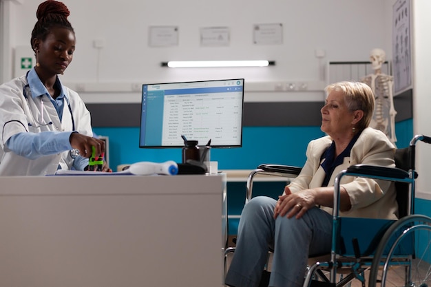 Mujer pensionista enferma en silla de ruedas explicando los síntomas de la enfermedad al médico terapeuta durante la cita médica en el consultorio del hospital. Médico médico estampar prescripción de tratamiento de medicamentos