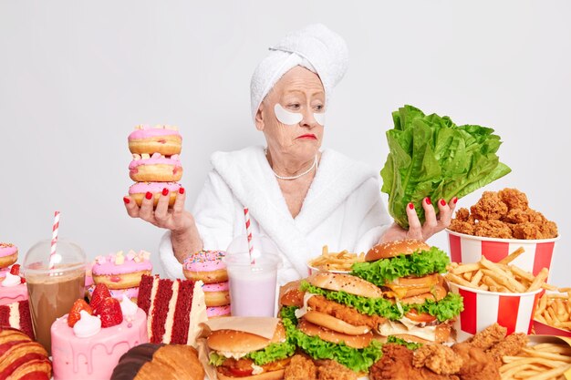 Mujer pensionista vacilante duda entre alimentos saludables y no saludables sostiene donas y vegetales verdes