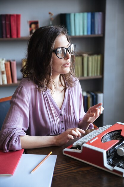 Mujer periodista en anteojos escribiendo en máquina de escribir en interiores