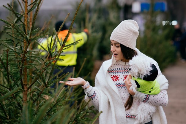 Mujer con un perro blanco en sus brazos cerca de un verde árboles de Navidad en el mercado