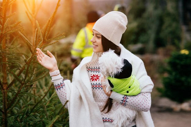 Mujer con un perro blanco en sus brazos cerca de un verde árboles de Navidad en el mercado