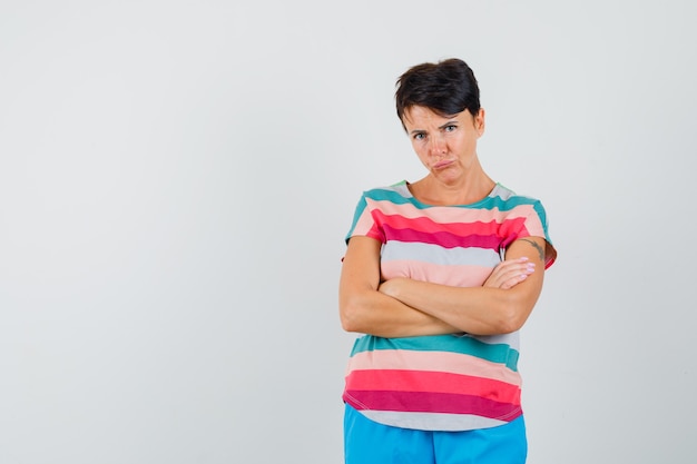 Mujer de pie con los brazos cruzados en camiseta a rayas, pantalones y mirada triste.