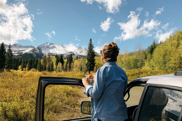 Mujer de pie cerca del coche disfrutando de la vista con árboles y montañas nevadas en la distancia