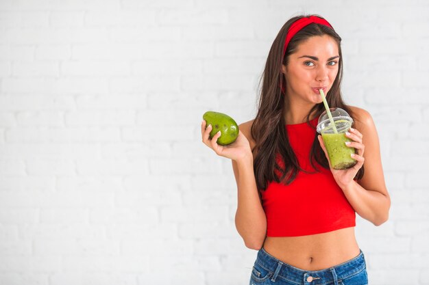 Mujer de pie contra la pared beber batidos de aguacate en vaso de plástico