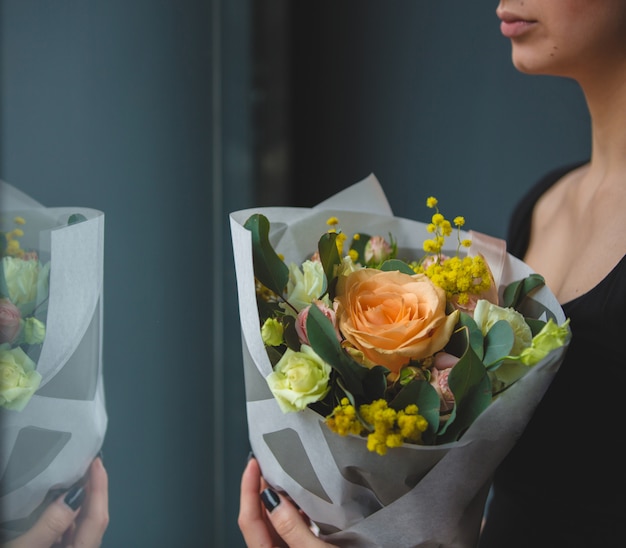 Una mujer de pie delante de una ventana y mirando hacia afuera con un ramo de rosas de coral en la mano