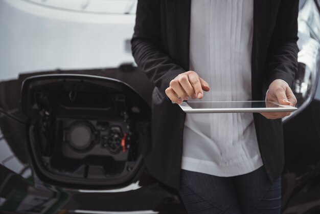 Mujer de pie junto al coche eléctrico y con tableta digital