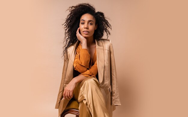 Mujer con piel bronceada con pelos rizados perfectos en elegante blusa naranja y pantalones de seda sentada en la pared beige de la silla vintage.