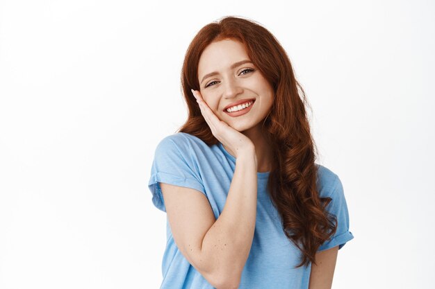 Mujer con piel natural fresca y limpia, cabello rojo, tocando la mejilla y sonriendo feliz y satisfecha, usando cosmética facial limpiadora para el cuidado de la piel, de pie sobre blanco
