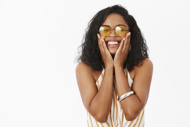 Mujer de piel oscura cerrando los ojos de alegría y placer sosteniendo las palmas en las mejillas, sonriendo ampliamente