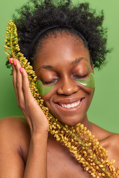 Mujer de piel oscura complacida mantiene los ojos cerrados sonríe ampliamente utiliza productos a base de hierbas naturales tiene cabello rizado