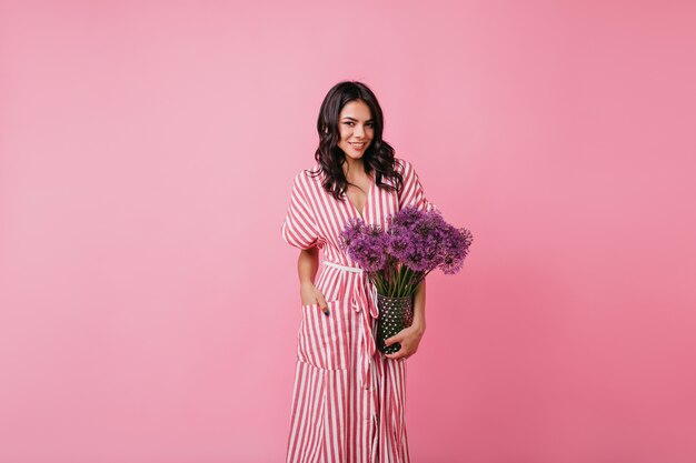 Mujer de piel oscura con confianza en la habitación rosa. Señora en vestido largo tiene jarrón de flores.