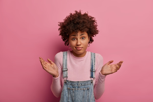 Mujer de piel oscura cuestionada insegura levanta las manos con expresión confusa, perpleja para dar una respuesta a una pregunta difícil, viste un atuendo casual, aislado sobre una pared rosa. Percepcion humana