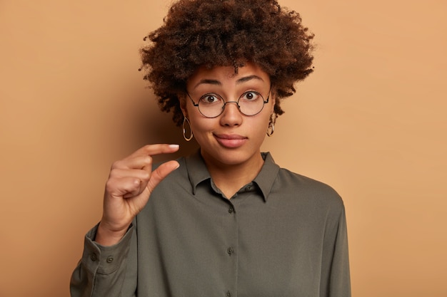 Mujer de piel oscura descontenta y disgustada da forma a un artículo de tamaño pequeño, confundida por algo muy pequeño, sonríe, usa lentes ópticos y camisa, explica y mide con los dedos, se para en el interior