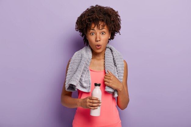 Mujer de piel oscura deshidratada conmocionada bebe agua después del entrenamiento, viste ropa de fitness y una toalla alrededor del cuello