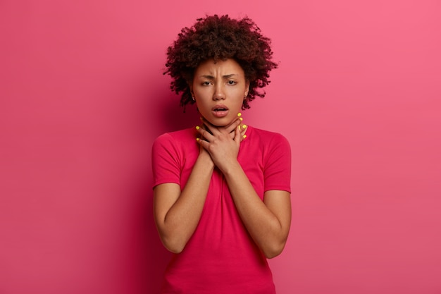 Mujer de piel oscura disgustada que tiene un ataque de asma, no puede respirar bien, mantiene las manos en el cuello, frunce el ceño, se asfixia o se ahoga, tose, tiene problemas de salud, usa ropa informal, aislado en rosa