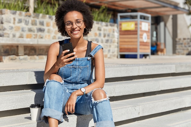 Mujer de piel oscura envía mensajes de texto por celular, chatea en redes sociales, viste petos andrajosos, se sienta en las escaleras, disfruta de café desechable, tiempo de recreación en la calle. Adolescente despreocupado con dispositivo