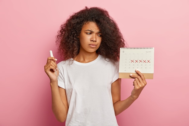Una mujer de piel oscura estresada y disgustada mira el calendario menstrual con cruces rojas marcadas.