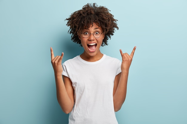 Mujer de piel oscura grita de felicidad, tiene la boca muy abierta, hace un gesto de rock n roll, usa un atuendo informal