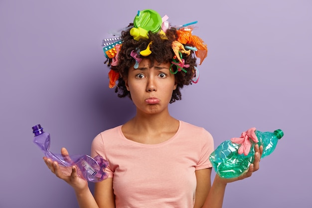 Mujer de piel oscura insatisfecha se preocupa por el medio ambiente limpio, sostiene dos botellas de plástico arrugadas, recoge basura por todas partes, está triste por los problemas de contaminación de la naturaleza, se preocupa por la ecología