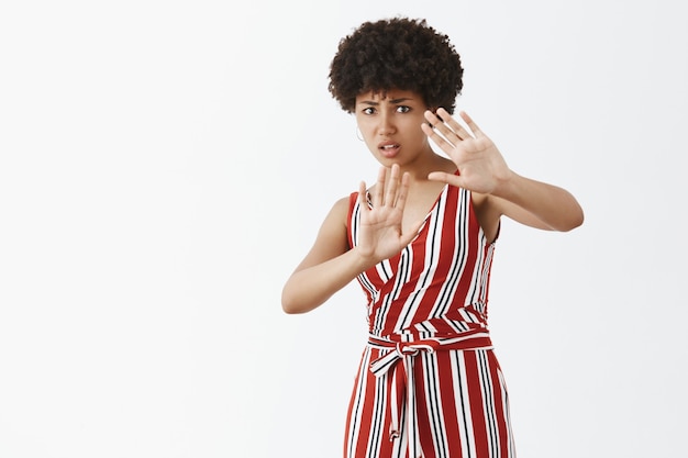 Mujer de piel oscura insegura, disgustada e infeliz con peinado afro en traje de moda levantando las palmas en gesto de protección, preocupado por derramar bebida en la ropa, frunciendo el ceño