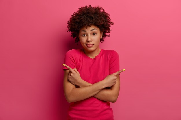 Una mujer de piel oscura interrogada toma una decisión, muestra dos formas, señala hacia los lados, tiene una expresión vacilante, usa una camiseta informal, posa sobre una pared rosada, mira con desconcierto o perplejidad.