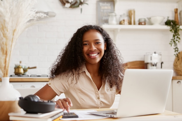 Mujer de piel oscura joven con estilo atractivo en camisa beige sentada en la mesa de la cocina, usando la computadora portátil, calculando el presupuesto, planeando las vacaciones, sonriendo felizmente. Mujer negra autónoma que trabaja desde casa