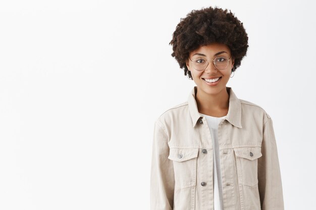 Mujer de piel oscura juvenil casual de aspecto agradable con peinado afro con gafas y camisa de moda sonriendo ampliamente y de pie sobre una pared gris con mirada despreocupada y relajada