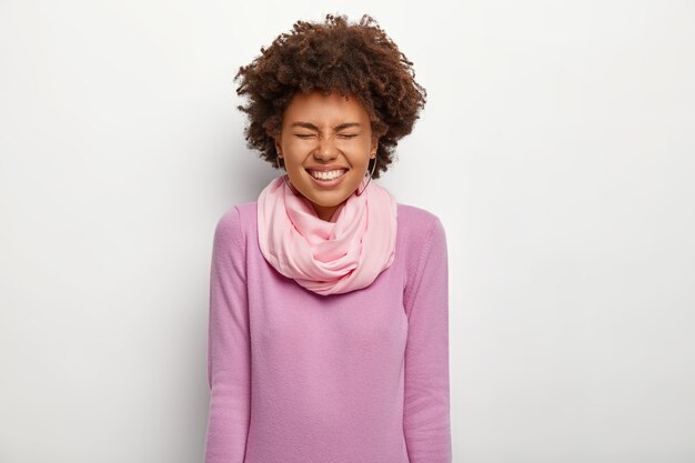 Mujer de piel oscura llena de alegría mantiene los ojos cerrados, se ríe feliz, viste un cuello alto de color púrpura con pañuelo de seda, tiene una amplia sonrisa