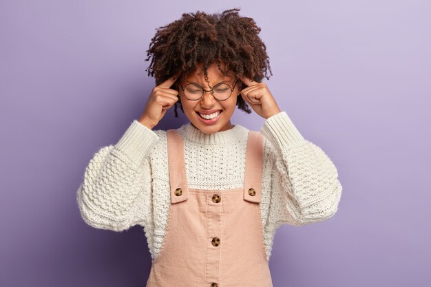 La mujer de piel oscura nerviosa maneja el estrés, alivia el dolor, mantiene los dedos índice en las sienes, sonríe, viste con un atuendo elegante, aislado sobre una pared púrpura. Sentimientos negativos y concepto de migraña.