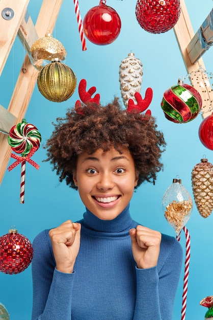 Mujer de piel oscura positiva con cabello rizado en cuello alto casual aprieta los puños anticipa el milagro se prepara para las vacaciones de Navidad vestida informalmente posa en el interior sobre juguetes de Año Nuevo. Evento festivo.