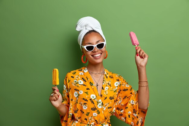 La mujer de piel oscura, positiva y despreocupada, sostiene un delicioso helado, paletas de helado, se divierte durante el verano, usa elegantes gafas de sol, una bata amarilla, una toalla envuelta en la cabeza, es golosa.