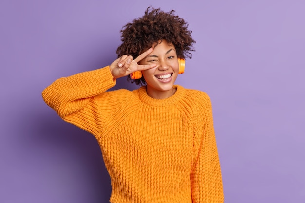 Mujer de piel oscura positiva hace gesto de paz sobre los ojos sonríe ampliamente se divierte se siente divertido escucha canciones agradables en poses de auriculares