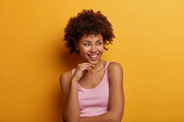 La mujer de piel oscura positiva mira hacia otro lado, ha satisfecho la expresión complacida, sostiene la mano debajo de la barbilla, habla casualmente, tiene una expresión soñadora, vestida con ropa casual, aislada en la pared amarilla.