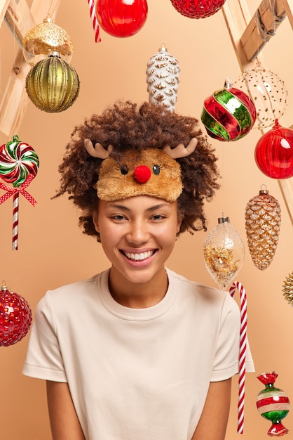 Mujer de piel oscura positiva usa antifaz para dormir de ciervo y camiseta casual sonríe ampliamente a la espera de poses de vacaciones de invierno