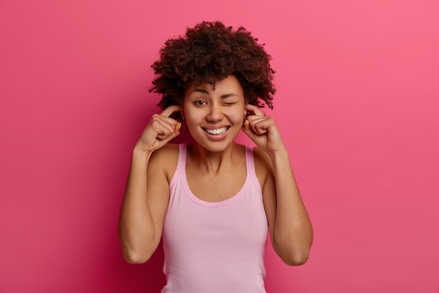 Mujer de piel oscura resentida y disgustada tapona los oídos con los dedos, ignora el sonido muy fuerte, frunce el ceño y muestra los dientes, no puede soportar el ruido, guiña el ojo, vestida de manera informal, aislada en la pared rosa