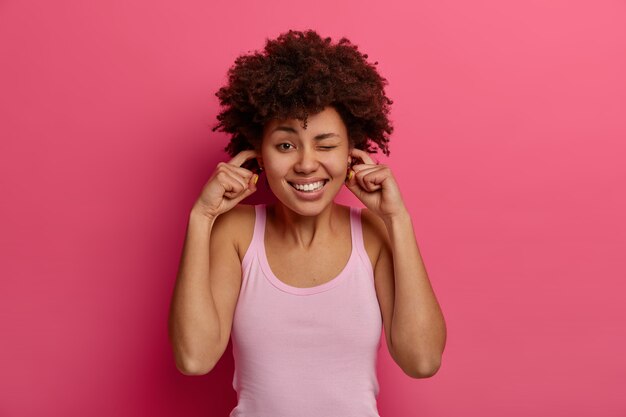 Mujer de piel oscura resentida y disgustada tapona los oídos con los dedos, ignora el sonido muy fuerte, frunce el ceño y muestra los dientes, no puede soportar el ruido, guiña el ojo, vestida de manera informal, aislada en la pared rosa