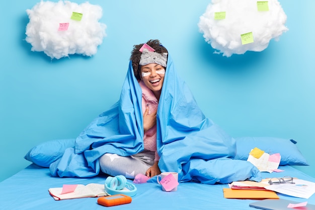 Foto gratuita mujer de piel oscura sincera positiva se ríe felizmente disfruta de un ambiente doméstico sereno envuelto en cálidas poses de manta en una cómoda cama rodeada de pegatinas de bloc de notas con ideas escritas sobre qué hacer