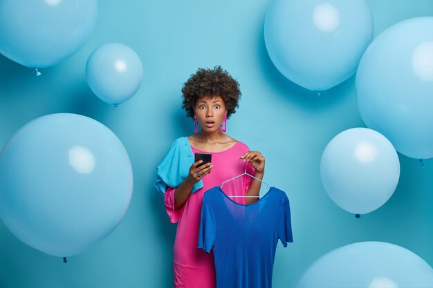 Mujer de piel oscura sorprendida elige el atuendo, sostiene el vestido azul en perchas, usa el teléfono inteligente y hace compras en línea en una boutique de moda, se prepara para una cita o una fiesta, aislado en una pared azul