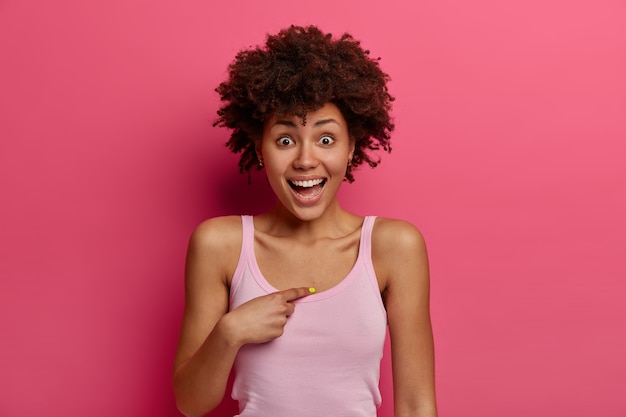 Una mujer de piel oscura sorprendida se señala a sí misma, fue ascendida o elegida, se ve con expresión feliz de sorpresa, tiene una mirada emocionada, usa ropa informal, está aislada en una pared rosa, no puede describir la felicidad