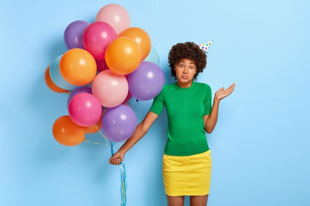 Mujer de piel oscura vacilante se siente confundida, levanta la palma de la mano, tiene peinado afro rizado, vestida con camiseta verde y falda amarilla, sostiene globos multicolores de colores y duda dónde celebrar cumpleaños